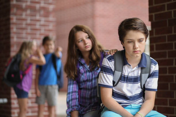 Friend comforts fellow student after he has been bullied outside the elementary school building,  Other students in background laugh at boy behind his back.
